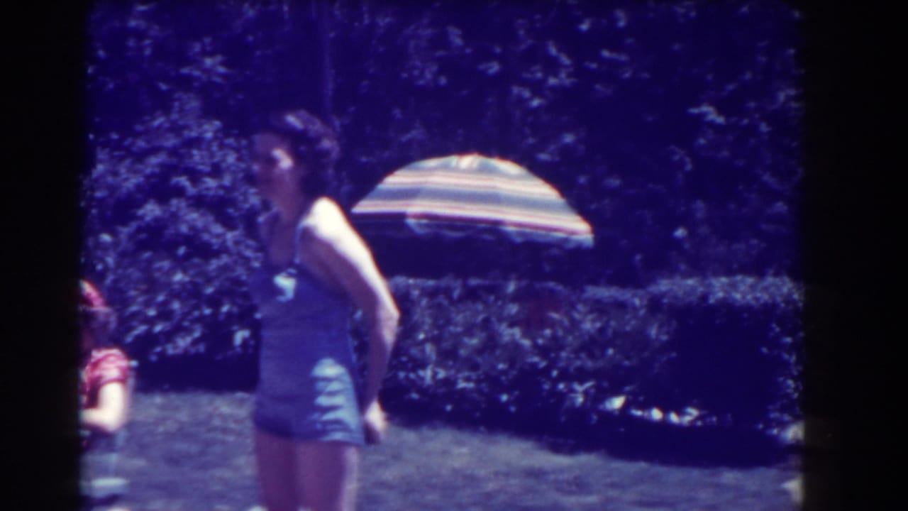 Child in a blue swimsuit stands on a lawn near a striped umbrella and hedges.