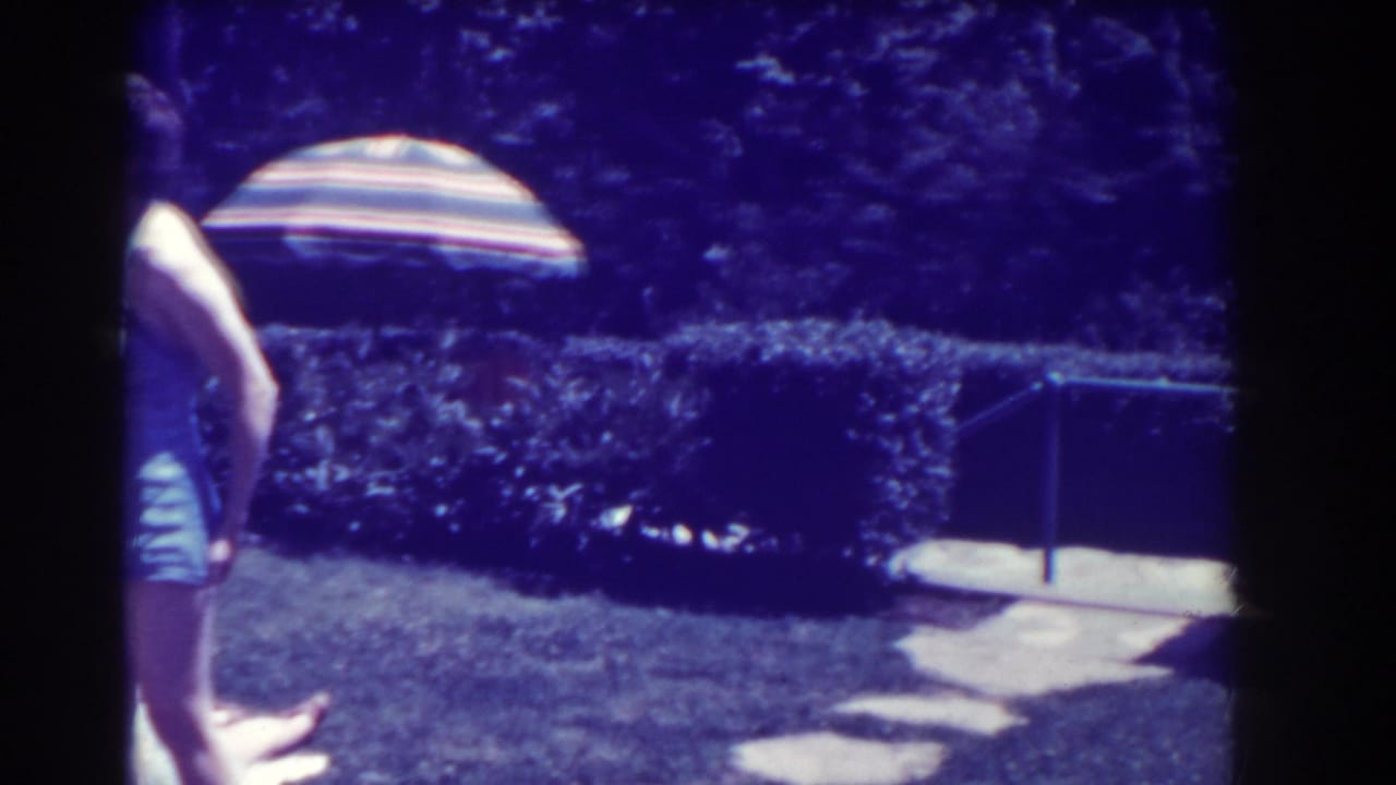 Child walks across a backyard lawn with hedges, a striped umbrella, and stone path nearby.