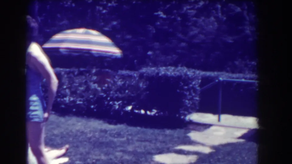 A child in a blue swimsuit moves across a Southampton backyard lawn, with a striped umbrella, hedges, and stepping stones visible.