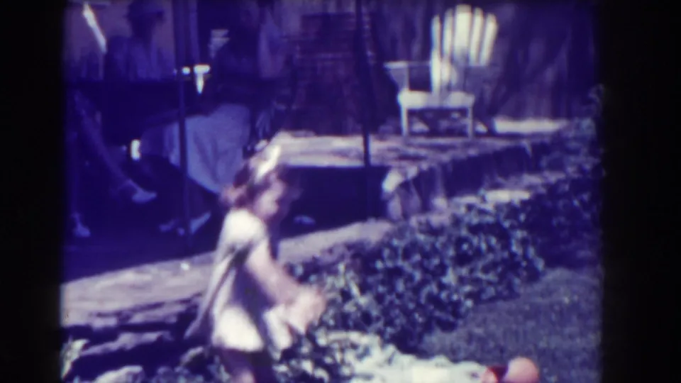 A home-movie frame shows an adult seated near a patio as a small child moves through a backyard with a lawn chair and planting bed.