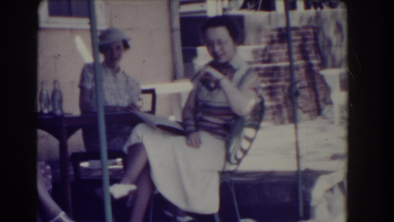 Three women sit on a shaded porch, one holding a book near a small table with bottles.
