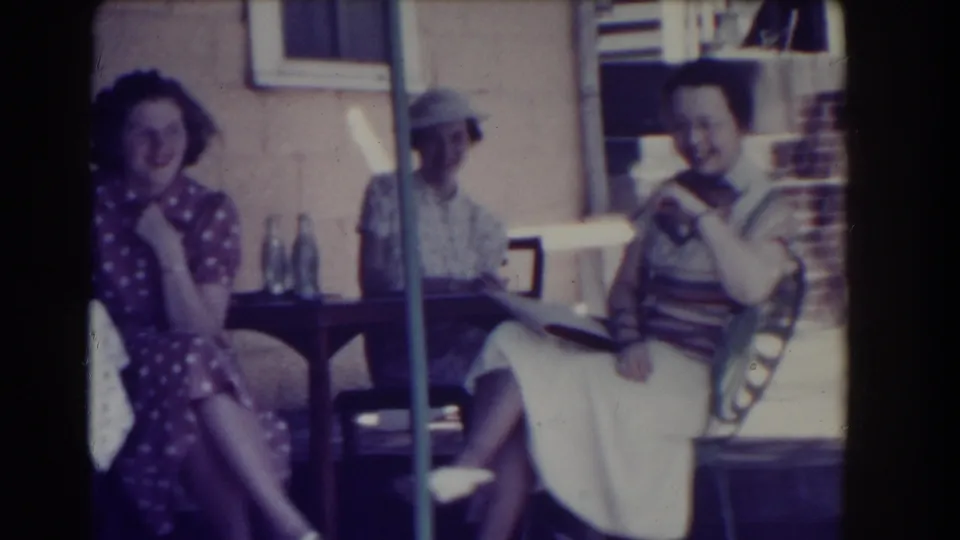 A woman in a polka-dot dress smiles toward the camera while two other women sit nearby on a 1937 Southhampton porch.