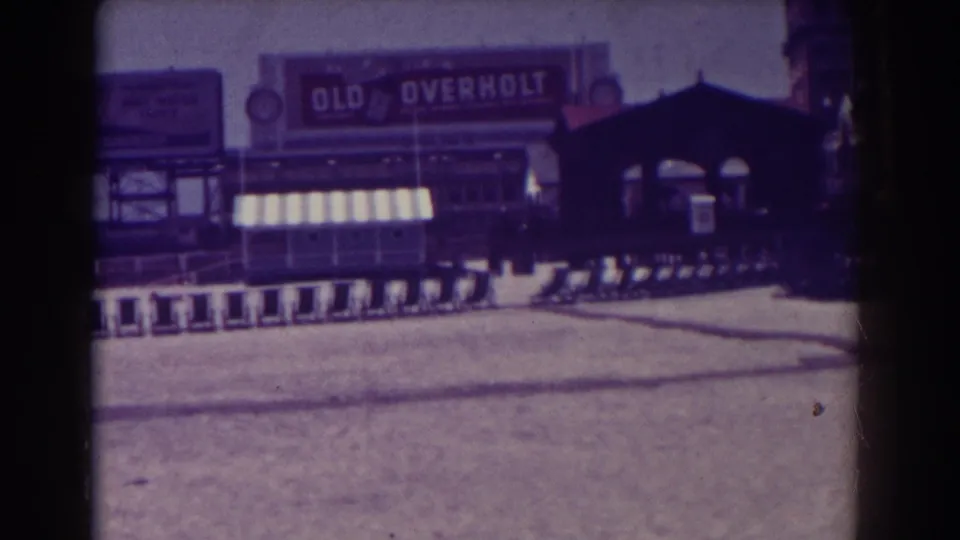 Wide 1938 Atlantic City boardwalk frame showing an Old Overholt sign, striped awnings, rows of chairs, a canopy, and distant pedestrians.
