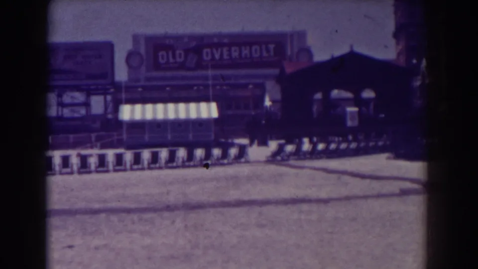 1938 Atlantic City home-movie frame showing training sleds on a sandy field below an Old Overholt billboard and striped awning.
