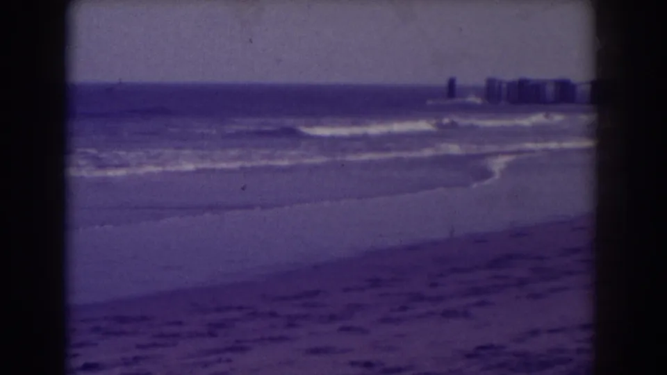 1938 Atlantic City beach view with surf, wet sand, horizon, and distant buildings captured in soft home-movie color.