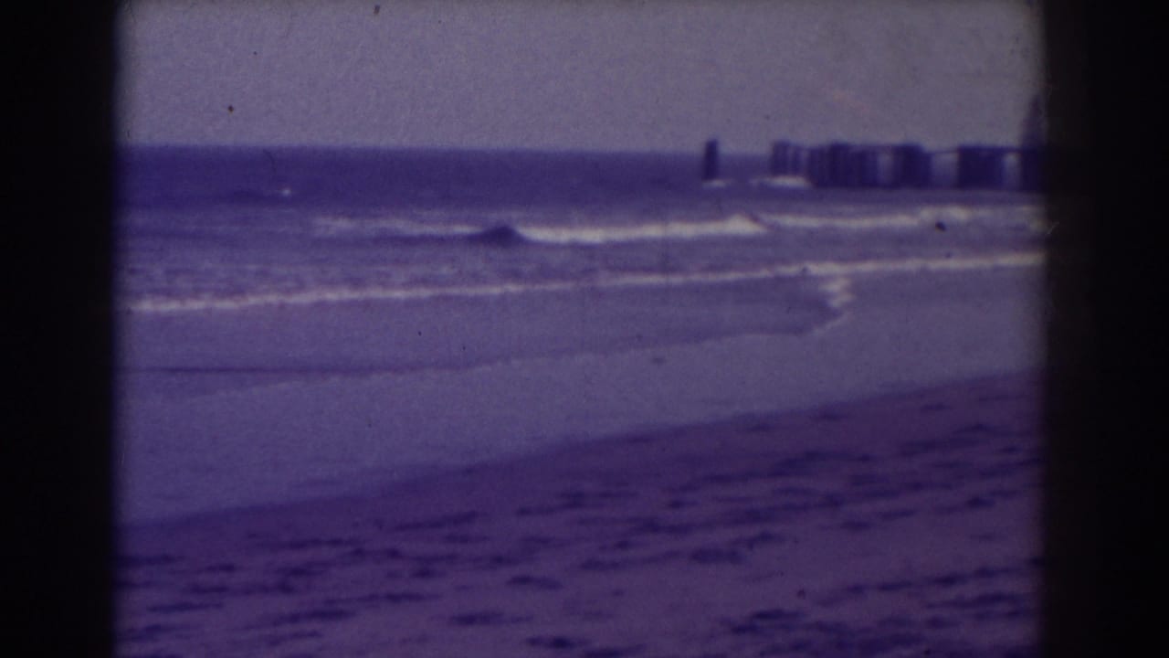 Purple-tinted beach and ocean surf with a distant pier on the horizon.