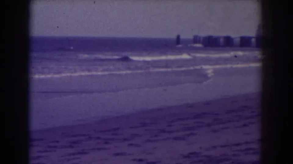 1938 Atlantic City home-movie frame showing a hazy beach, rolling surf, and a distant pier through 8mm film grain.