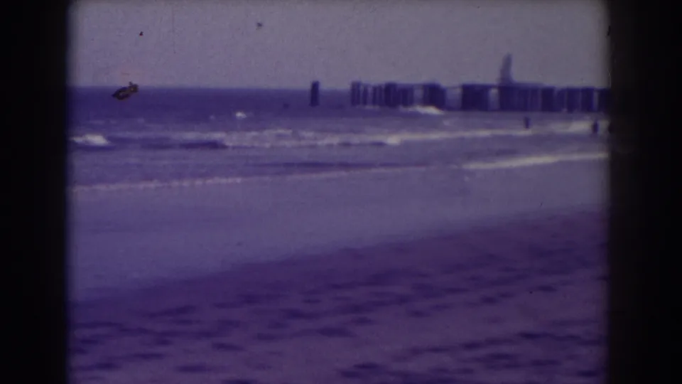 1938 Atlantic City shoreline view with wet sand, low waves, and a distant pier softened by home-movie film texture.