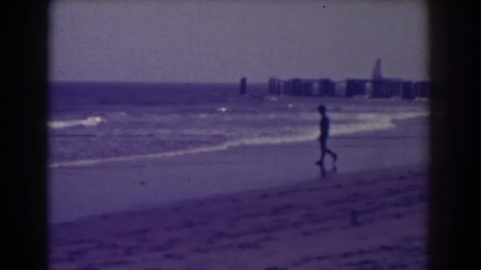 Atlantic City shoreline view with a lone walker near the waterline, distant pier forms, and purple-tinted 8mm film texture.