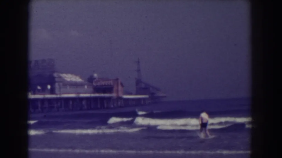 A beachgoer in a light shirt and dark shorts moves through shallow surf near Atlantic City pier buildings and signs.