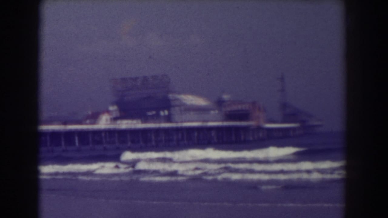 Soft 8mm view of waves breaking below a long pier with buildings at the far end.