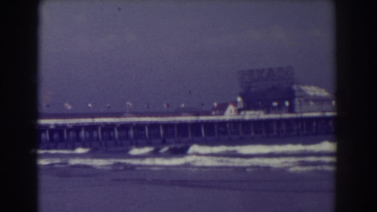 Long pier with flags above rolling surf, seen through soft faded 8mm film.