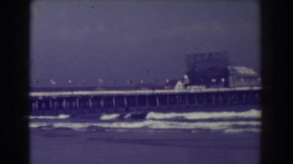 1938 Atlantic City frame with a long pier, small flags, foamy surf, hazy sky, and structures at the pier end.
