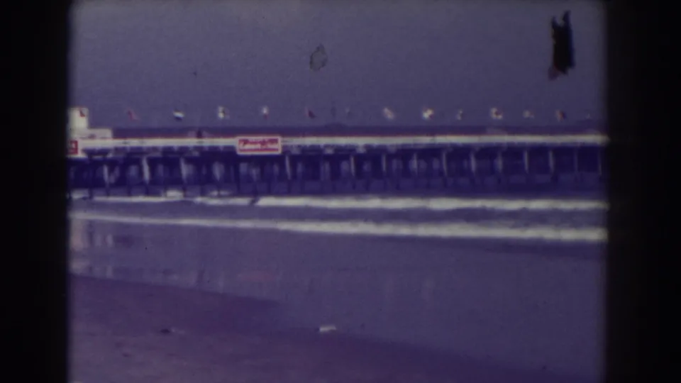 1938 Atlantic City home-movie frame showing a long pier beyond surf and wet sand, with flags and visible signage along the structure.