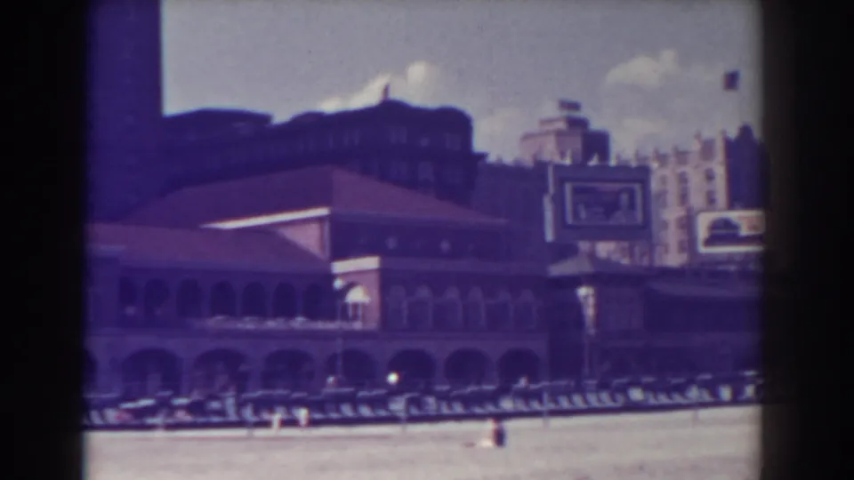 Atlantic City boardwalk buildings appear across the sand, with arched lower levels, taller structures behind, and small distant figures near the shorefront.