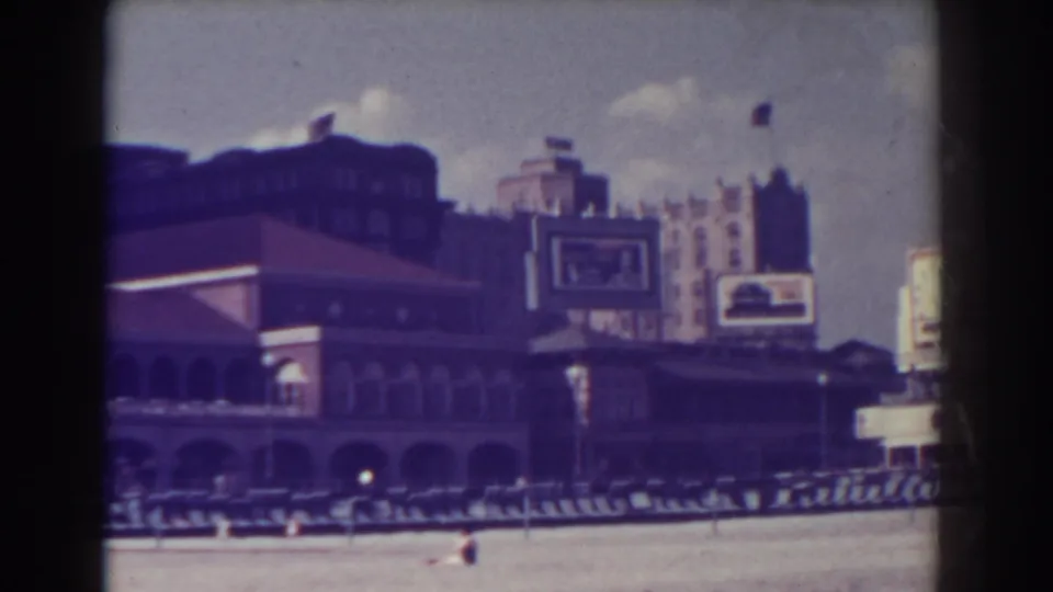 A visitor-shot 8mm frame shows Atlantic City beachfront architecture with arched openings, boardwalk railings, sand, and several distant people.