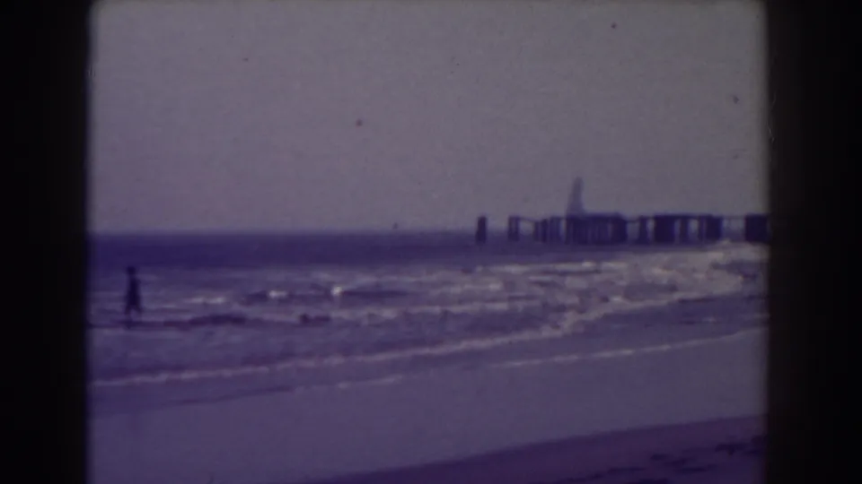 Blue-toned 8mm view of an Atlantic City beach, with one distant figure and a pier beyond the surf.