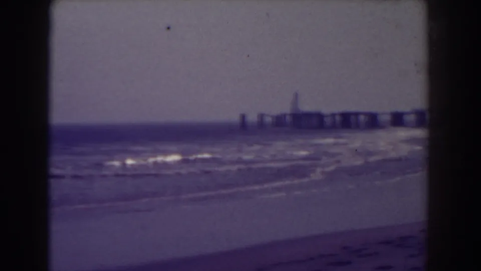 1938 Atlantic City beach view with low surf, a distant pier, soft focus, purple color cast, and dark 8mm frame edges.