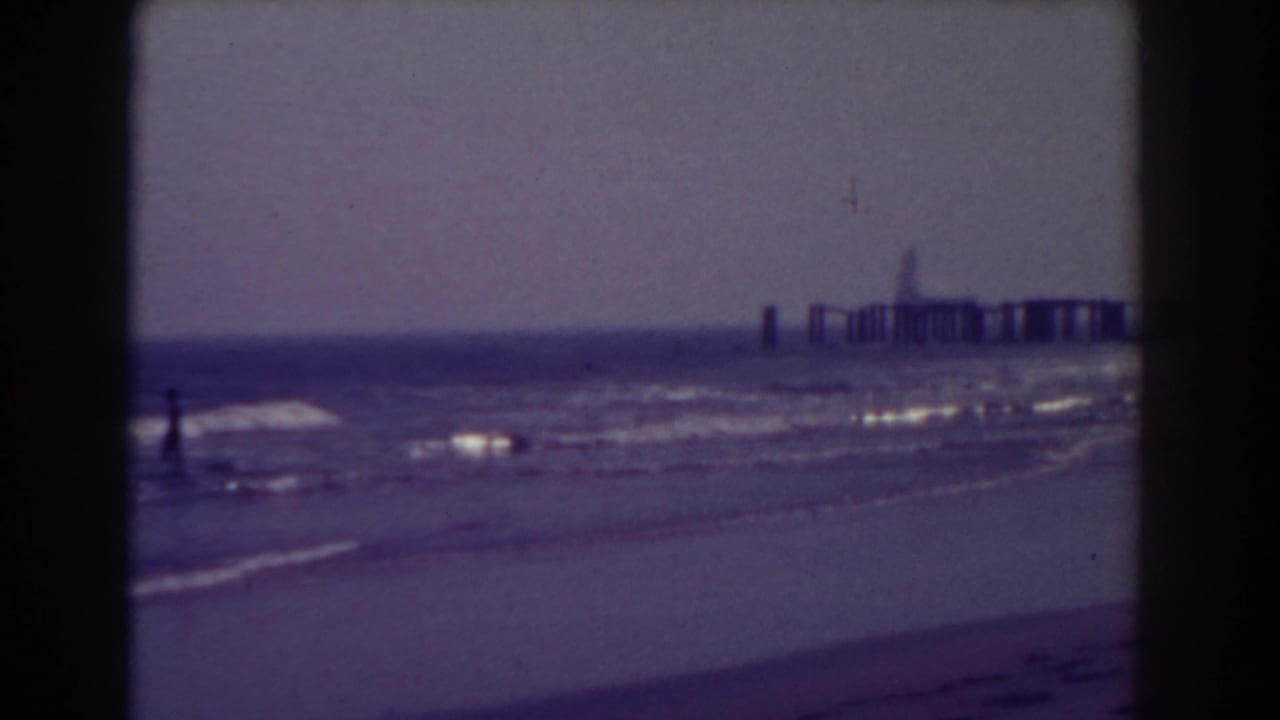 A small figure stands near the surf with a distant pier on the horizon.