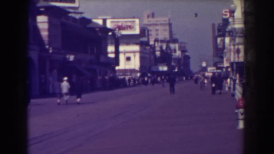 1938 Atlantic City street view with pedestrians walking past storefronts and signs in soft 8mm home-movie color.