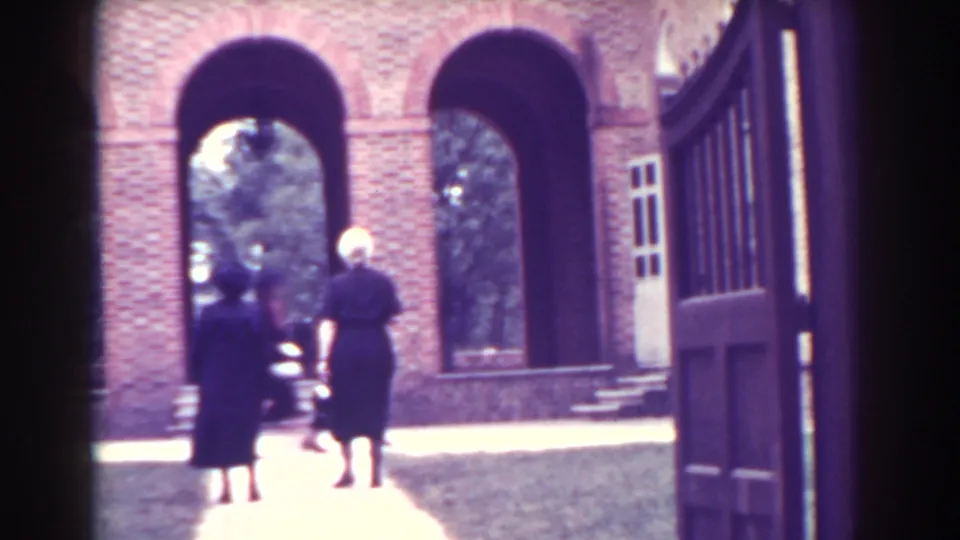 1938 Atlantic City home-movie frame with two women walking near brick arches, steps, trees, and an open paneled gate.