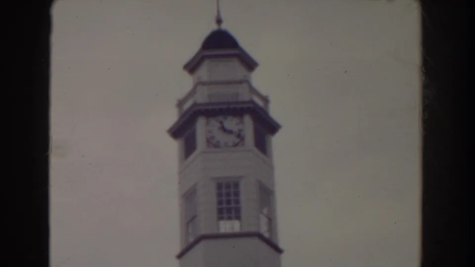 1938 Atlantic City home-movie frame centering the white clock tower, dark cap, clock face, and tower window against a washed pale sky.
