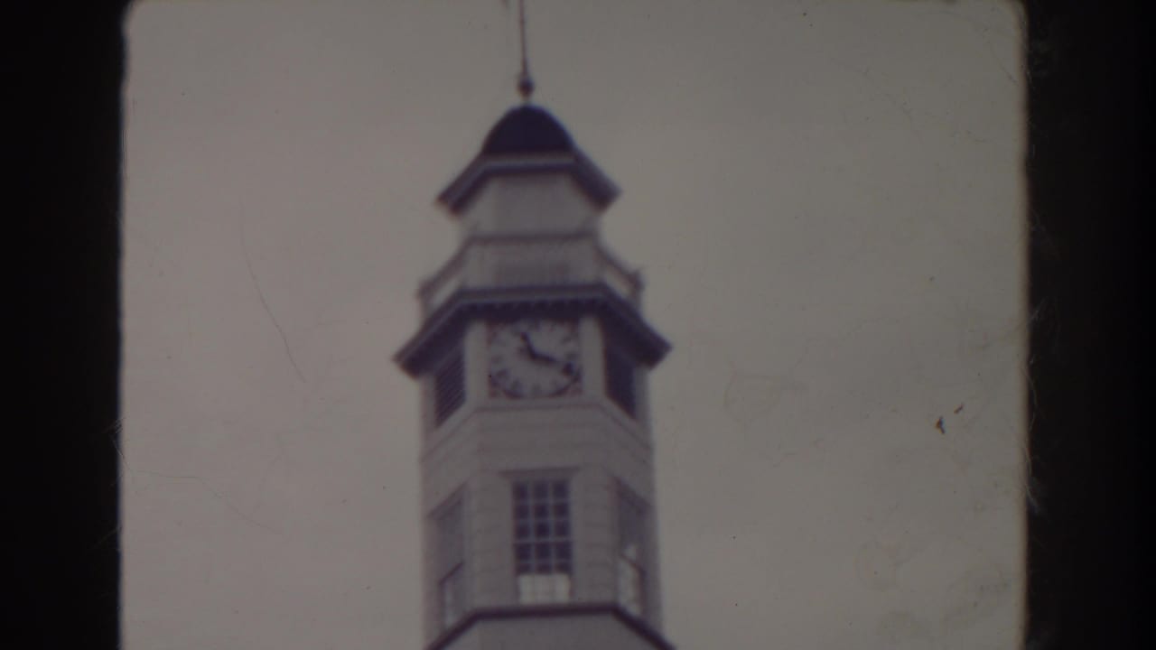 Soft 8mm view of a clock tower with a flag above it against a pale sky.