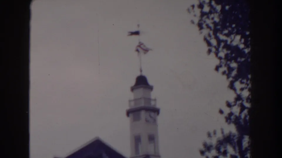 Atlantic City home-movie frame showing the upper clock tower with two flags flying above it and tree branches framing the right edge.