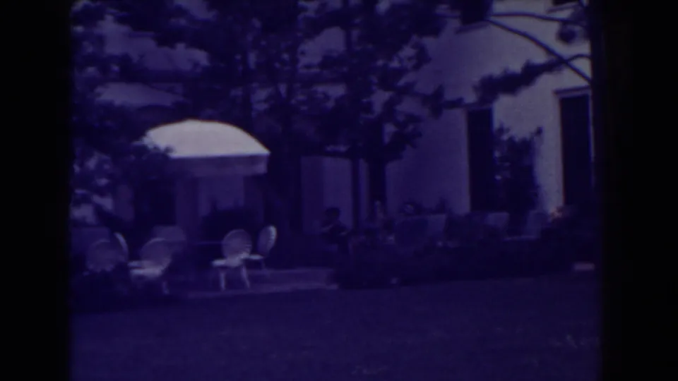 1938 Atlantic City home-movie frame showing a backyard patio with white umbrella, lawn chairs, trees, and distant seated people.
