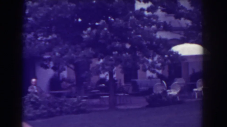 Atlantic City 1938 home-movie frame with a backyard lawn, white patio umbrella, chairs, trees, shrubs, and distant seated people.