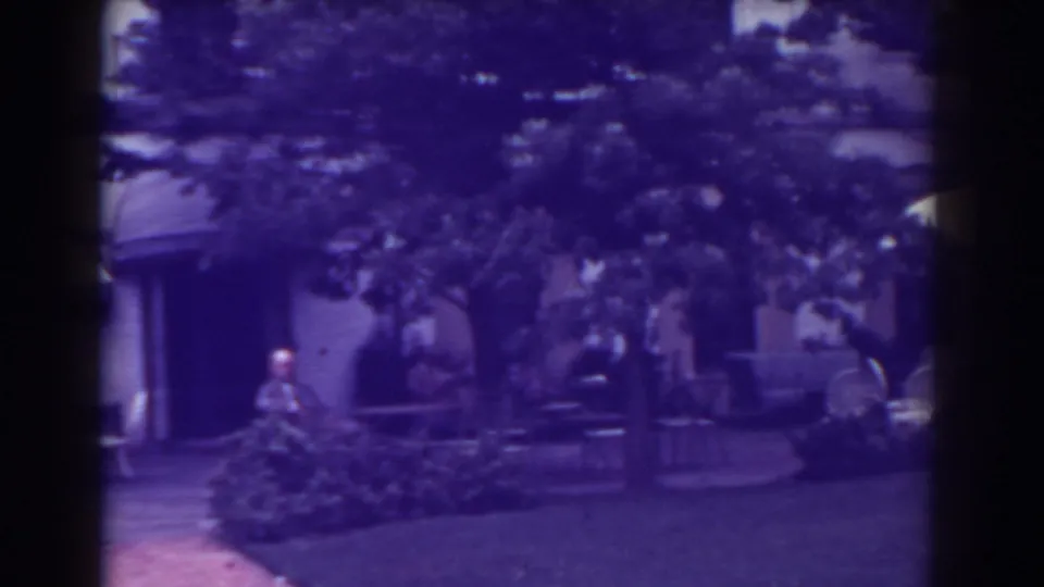 1938 Atlantic City home-movie frame showing a courtyard facade with awnings, outdoor chairs, shrubs, trees, and a distant standing man.