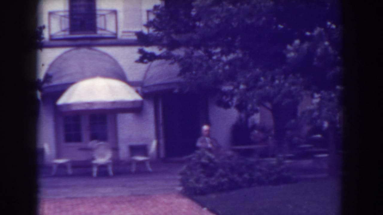 Building entrance with rounded awnings, white chairs, small tables, trees, shrubs, and a man at right.
