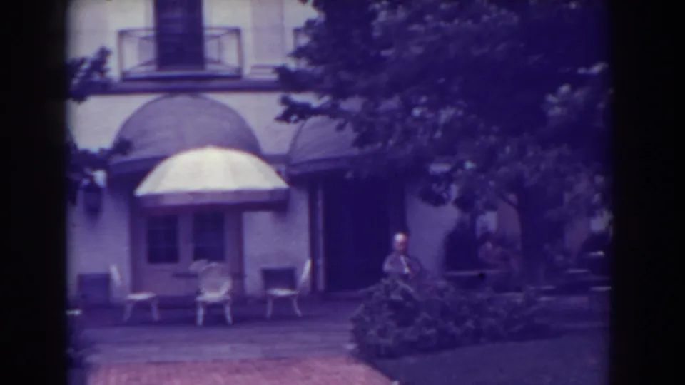 Closer 1938 Atlantic City frame of an exterior entrance with a large awning, patio chairs, shrubs, and a distant standing man.