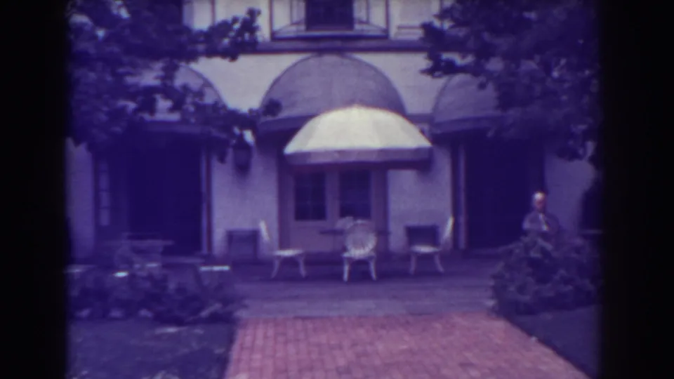 1938 Atlantic City courtyard frame with a brick walkway, arched awnings, outdoor seating, landscaped shrubs, and a distant man.