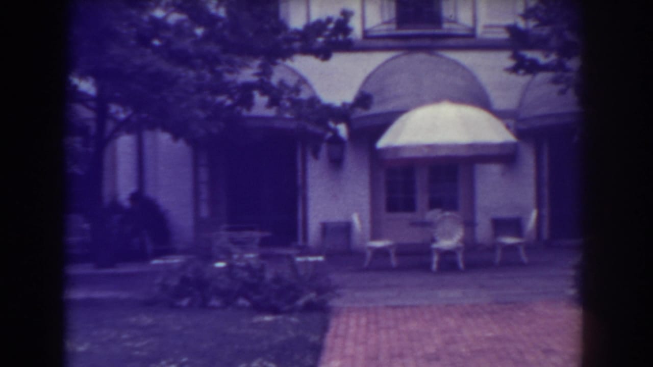Patio chairs and a light awning outside a large building, partly framed by trees.