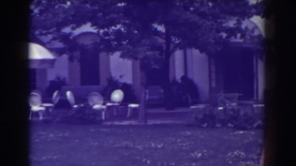 1938 Atlantic City frame showing several white patio chairs, a brick path, lawn, trees, and the shaded facade of a large building.