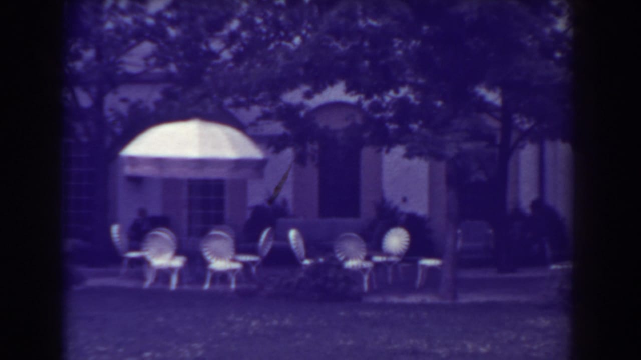 Patio chairs and tables on a lawn beside a pale building with awnings and trees.