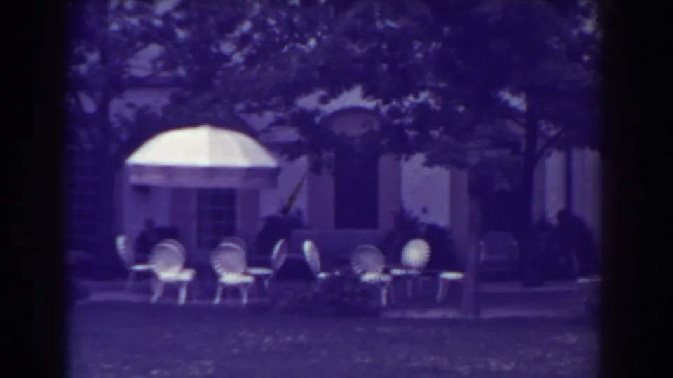 1938 Atlantic City home-movie frame showing outdoor patio seating, white awnings, lawn, and a pale building partly shaded by trees.