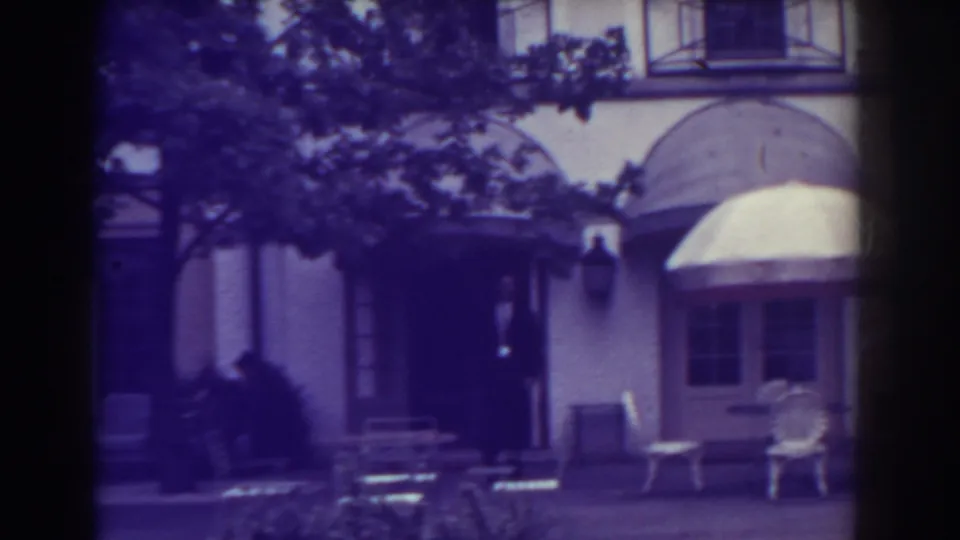 1938 Atlantic City home-movie frame showing a dark-suited adult near the entrance, with awnings and patio furniture in view.