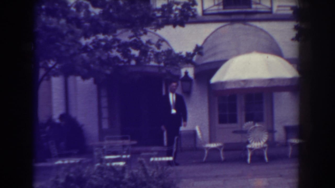 Man in a dark suit stands near a doorway beside patio chairs and a large umbrella.