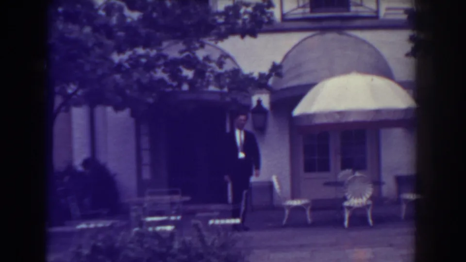 1938 Atlantic City home-movie frame showing a suited man near a courtyard doorway with white chairs, umbrella, awnings, and trees.