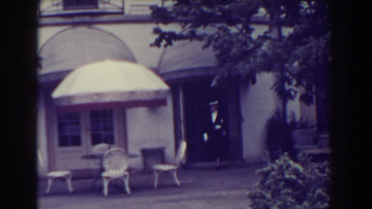 Man in a dark suit and hat steps out near a doorway beside courtyard furniture.