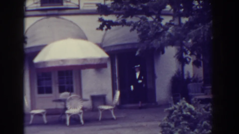 1938 Atlantic City frame with a well-dressed man near a building entrance, patio chairs, umbrella, shrubs, and arched awnings.