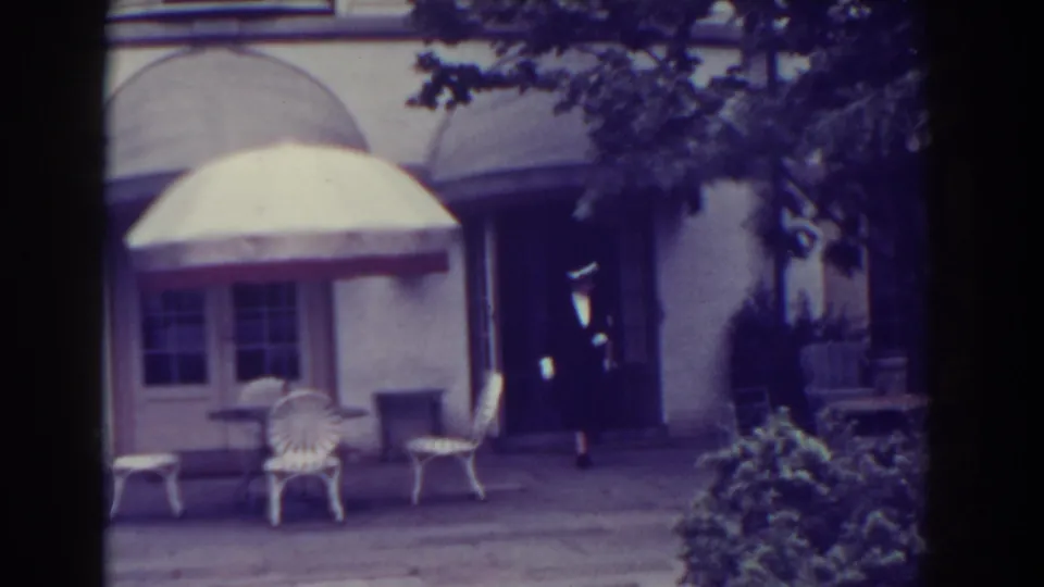 1938 Atlantic City home-movie frame showing a suited man walking past white patio chairs and a large umbrella outside a building.