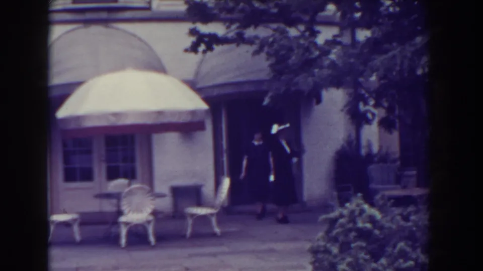1938 Atlantic City frame showing two formally dressed figures near an exterior doorway, with white chairs, umbrella, shrubs, and awnings visible.