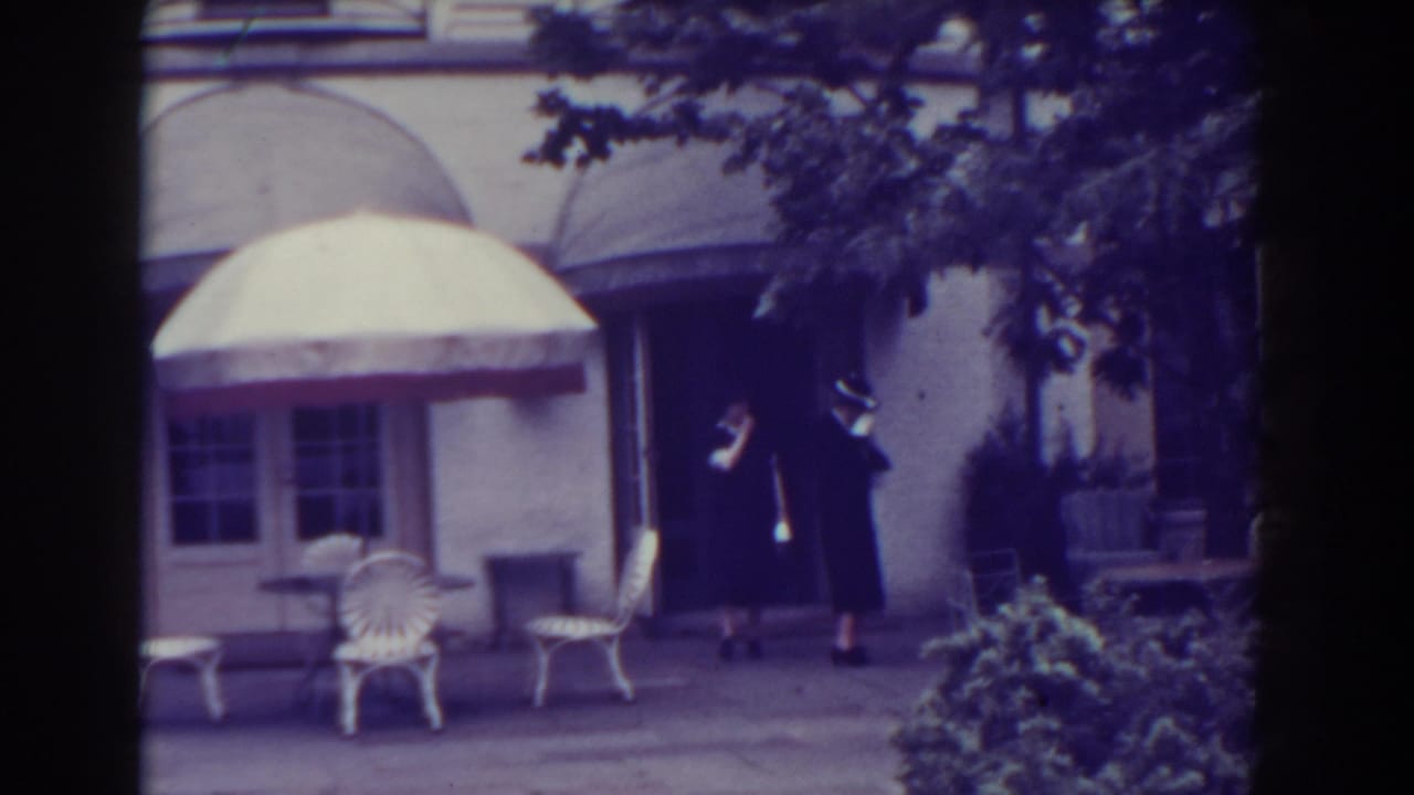Two women stand or begin walking outside a building with white patio chairs and an umbrella.