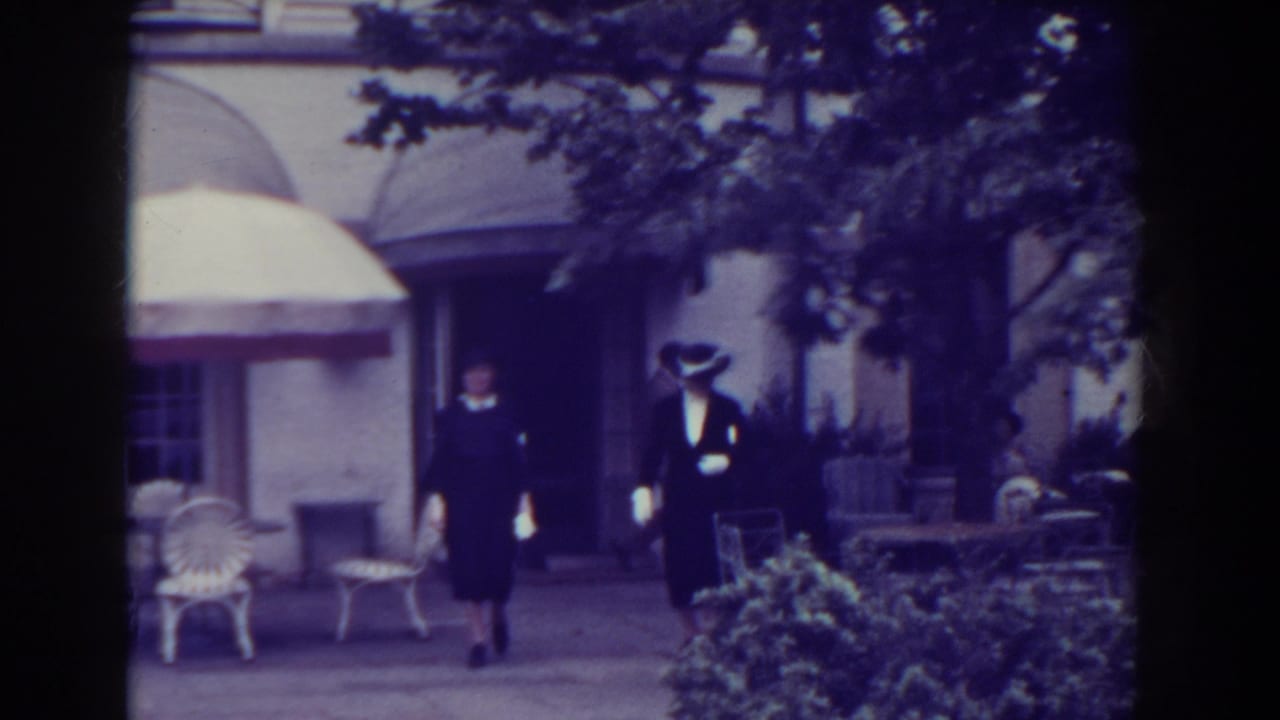 Two women in 1930s attire walk along a paved path in Atlantic City, with white chairs, an umbrella, shrubs, and a building behind them.