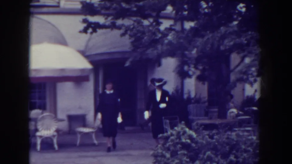 Two women in 1930s attire walk along a paved path in Atlantic City, with white chairs, an umbrella, shrubs, and a building behind them.