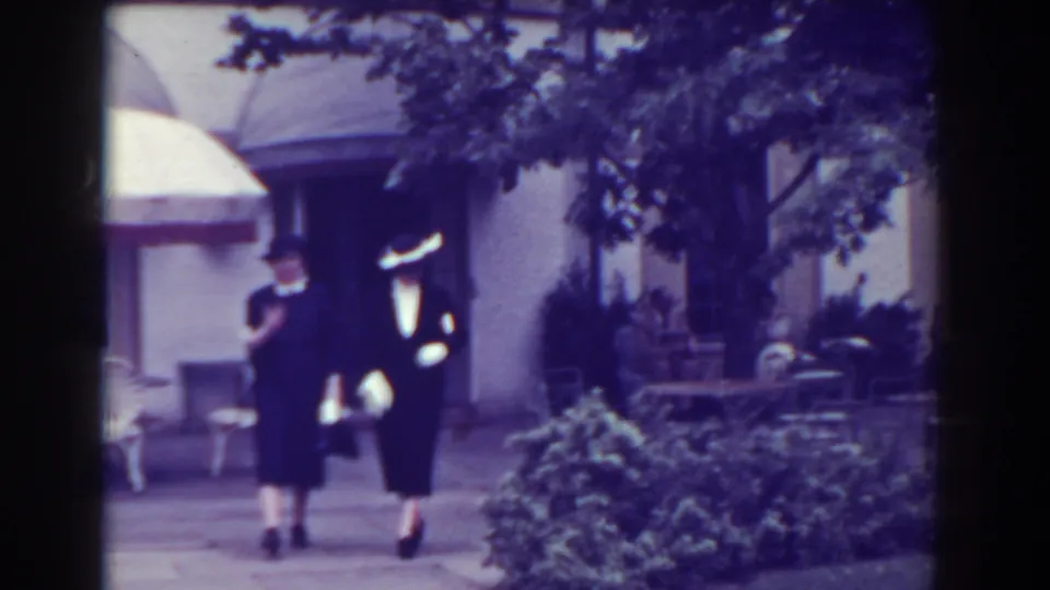 Near the end of the 1938 Atlantic City clip, two women walk close together past shrubs, chairs, and a shaded building entrance.
