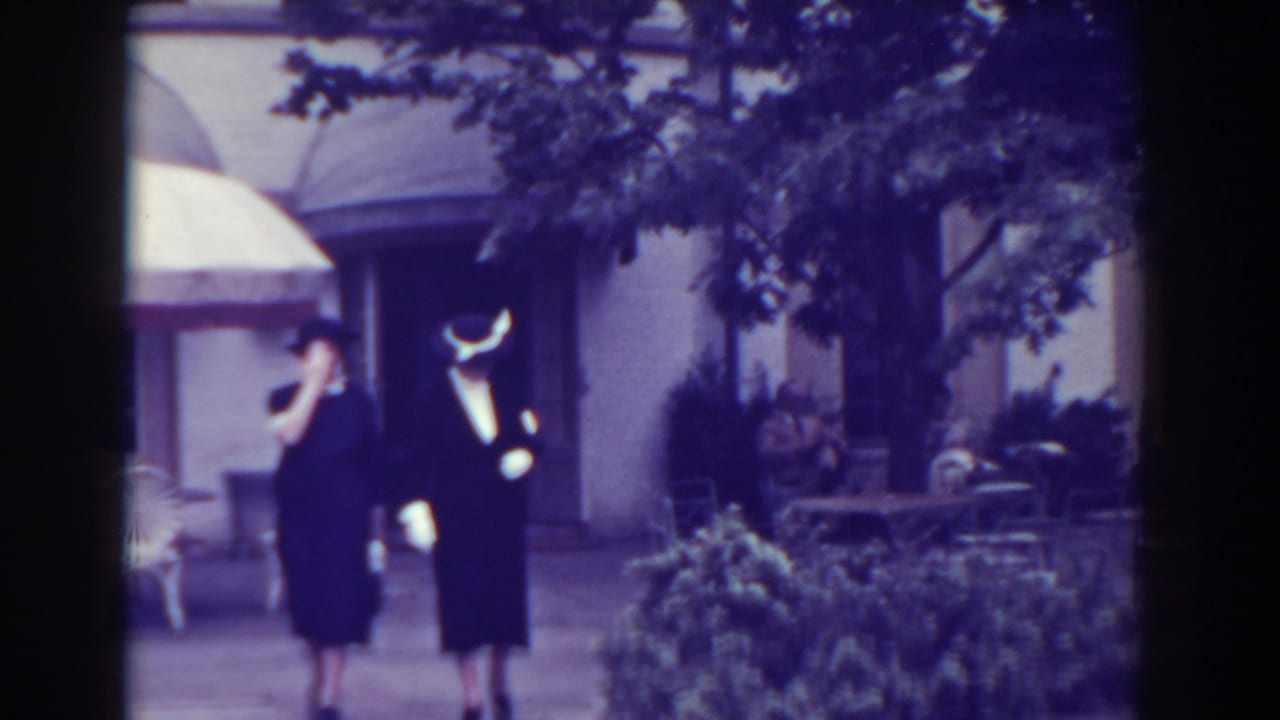 Two women in dark dresses and hats walk outdoors near shrubs and a building entrance.